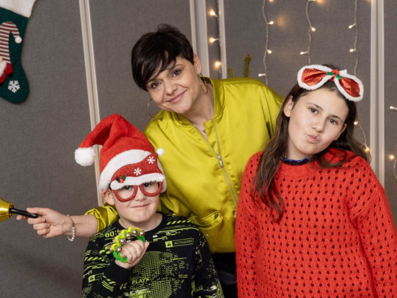 Kobieta w żółtej kurtce i dwoje dzieci pozuje po nagraniach w świątecznym studiu; jedno dziecko trzyma dzwonek, drugie ma czapkę Mikołaja i zabawne okulary