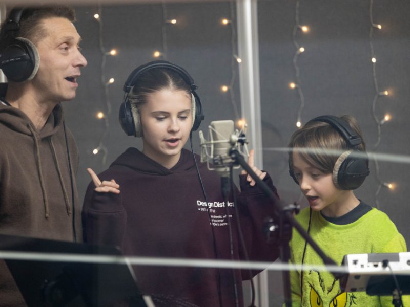 Ojciec i dwoje dzieci stoją przy mikrofonie w studio nagraniowym, wszyscy w słuchawkach, śpiewają świąteczną piosenkę