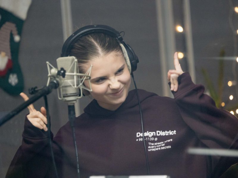 Nastolatka w ciemnej bluzy pozuje przy mikrofonie w studio, uśmiecha się i pokazuje gest taneczny