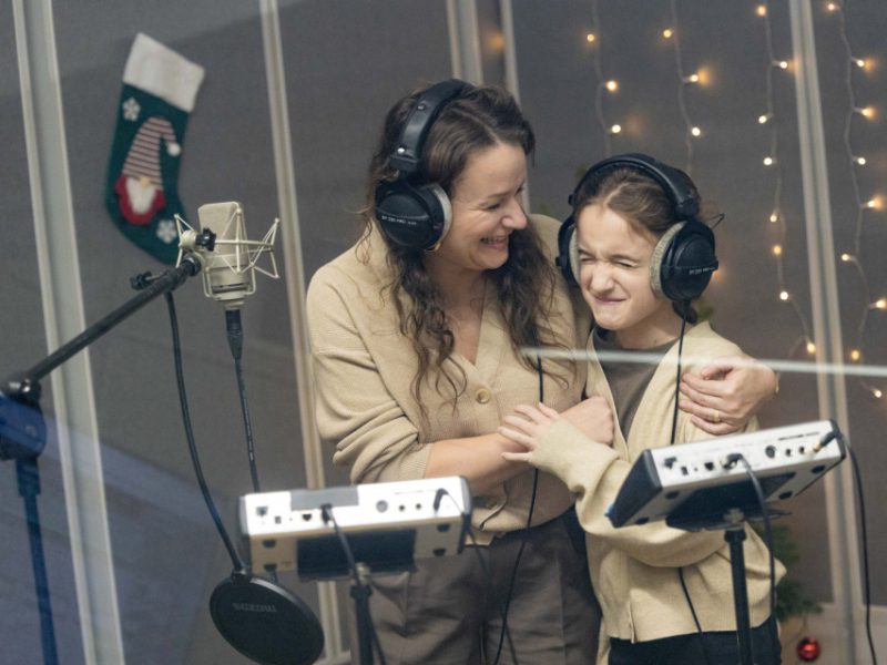 Mama przytula córkę w studio nagraniowym; obie mają na sobie słuchawki i śmieją się podczas nagrań
