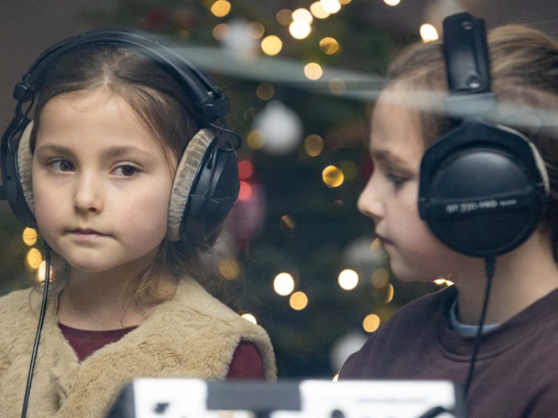 Dziewczynka w futerkowej kamizelce stoi w studio i patrzy przed siebie, obok niej inne dziecko w słuchawkach