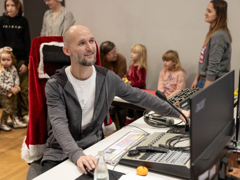 Mężczyzna siedzi przy stole realizatora dźwięku, obsługując sprzęt w studiu nagraniowym. Za nim stoją dzieci i dorośli czekający na nagranie