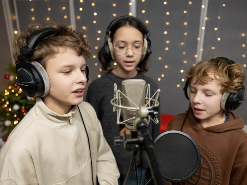 Troje dzieci śpiewa wspólnie przy mikrofonie w studiu nagraniowym, wszyscy mają na głowie duże słuchawki