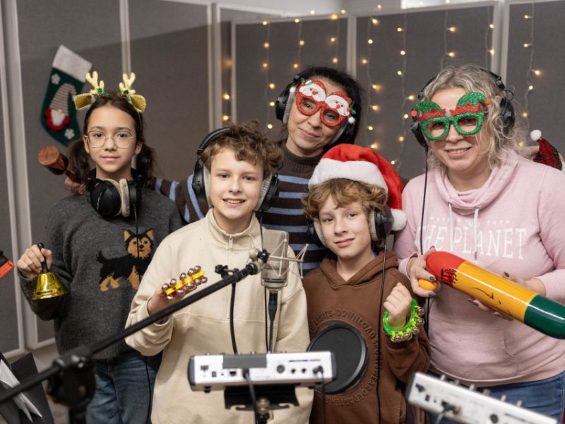 Grupa dzieci i dwóch dorosłych osób pozuje z instrumentami perkusyjnymi w świątecznym studiu nagraniowym. Wszyscy mają słuchawki i świąteczne akcenty w ubraniach