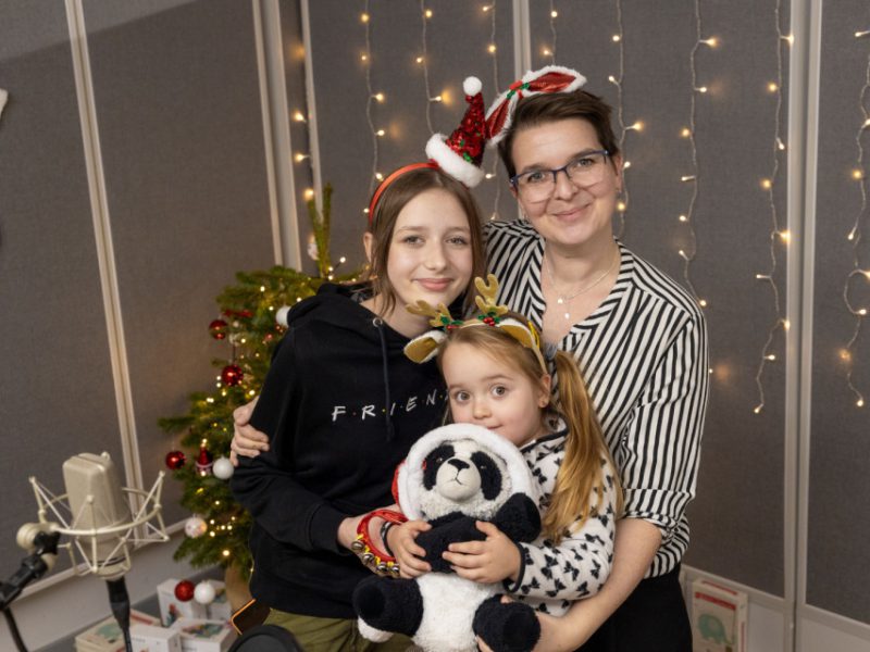 Dorosła kobieta i dwie dziewczynki stoją razem w studiu nagraniowym przed choinką. Wszystkie mają świąteczne opaski, a młodsze dziecko trzyma pluszową pandę