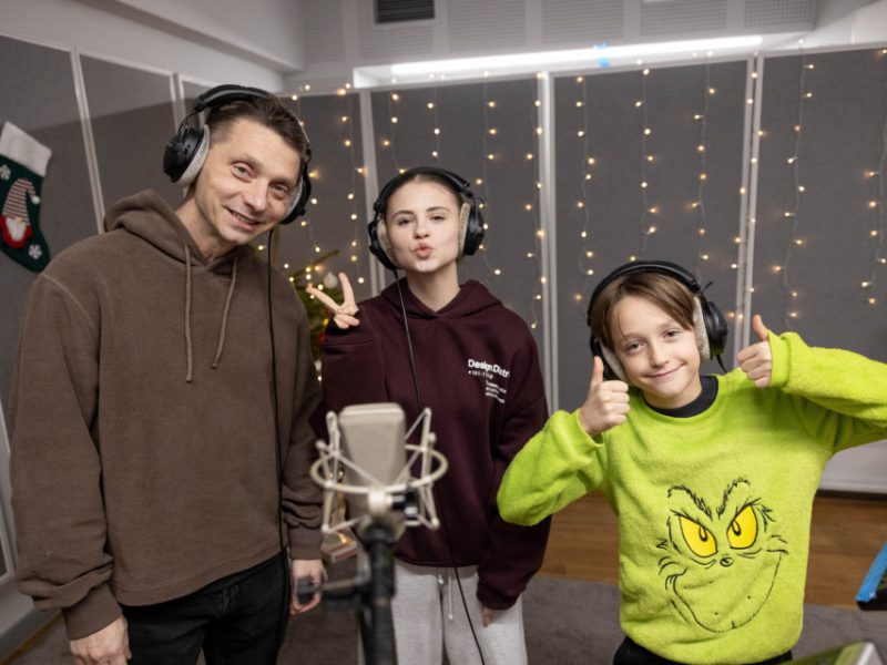 Ojciec i dwoje dzieci pozują przed mikrofonem w studio; syn pokazuje podniesione kciuki, a córka robi znak pokoju