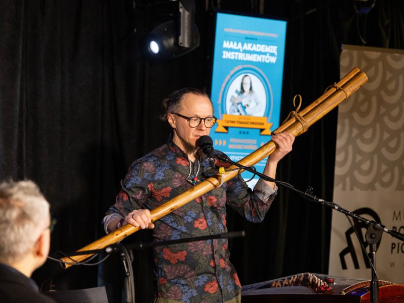 Muzyk przy mikrofonie trzyma długi drewniany instrument (didgeridoo) i opowiada o nim; w tle ekran „Mała Akademia Instrumentów” oraz baner muzeum