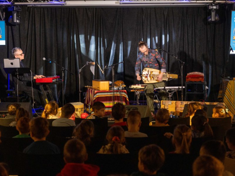 Szeroki plan sceny: dwóch muzyków i zestaw instrumentów, przed sceną siedzące dzieci; po bokach ekrany z grafiką wydarzenia