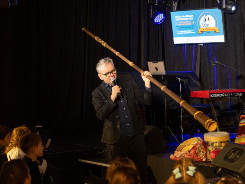 Prowadzący z mikrofonem prezentuje długi drewniany instrument dęty (didgeridoo); dzieci obserwują z pierwszych rzędów, w tle ekran z napisem „Mała Akademia Instrumentów”