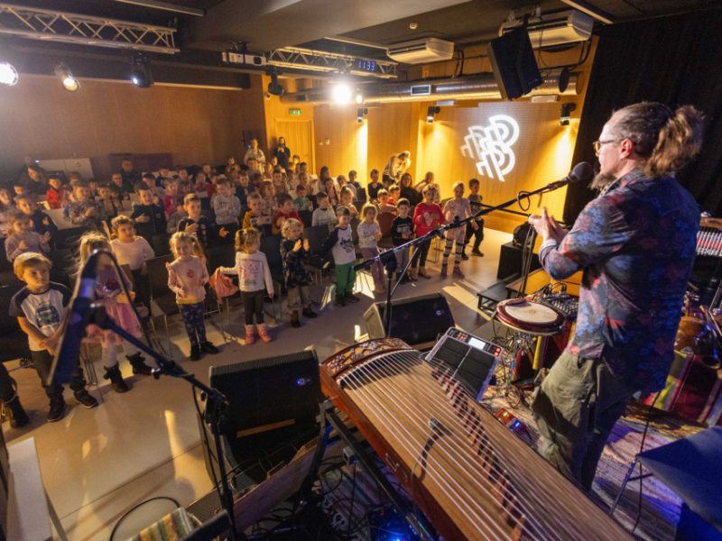 Muzyk przy mikrofonie prowadzi warsztaty na scenie; przed nim grupa dzieci, w tle sala i oświetlenie sceniczne