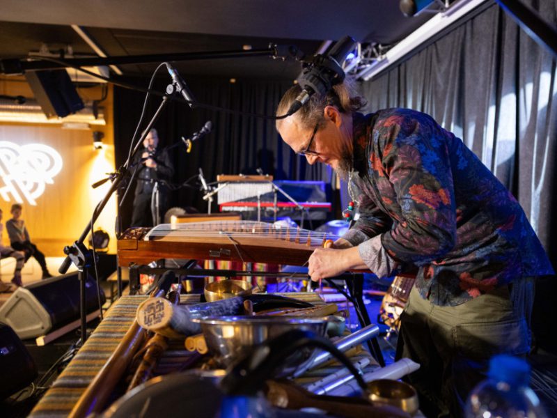 Muzyk pochylony nad instrumentem strunowym na scenie; na stole rozłożone instrumenty i rekwizyty, w tle widownia