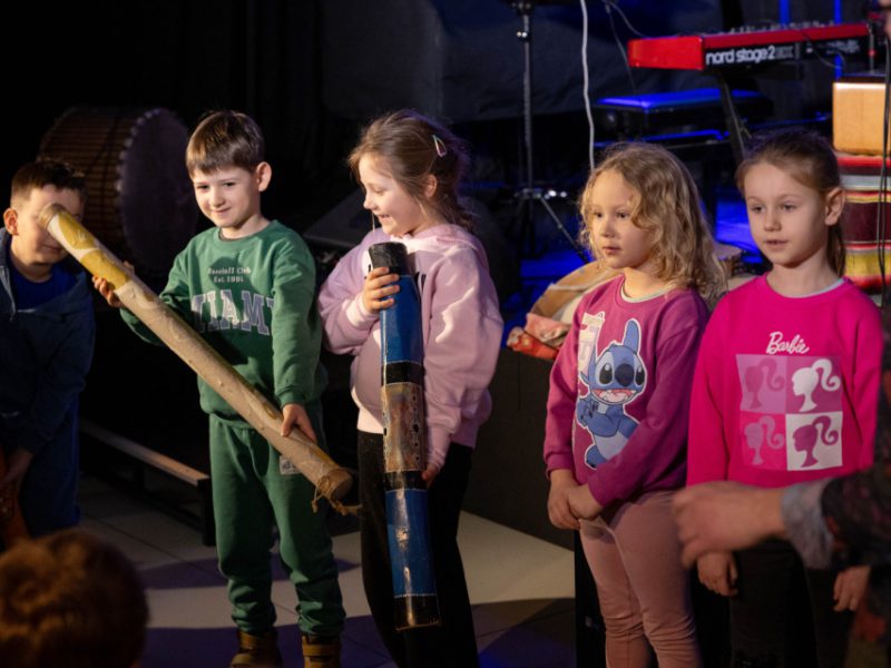 Dzieci na scenie trzymają długie drewniane rurki i instrumenty perkusyjne podczas warsztatów muzycznych