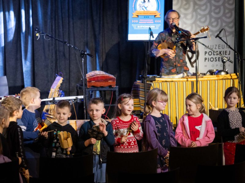 Grupa dzieci stoi z przodu sceny podczas warsztatów muzycznych; w tle prowadzący przy mikrofonie i zestaw instrumentów