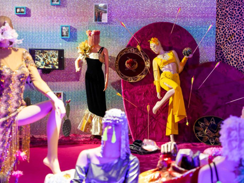 Scena z wystawy przedstawiającej manekiny w barwnych, teatralnych strojach; jeden stoi na tle sercowatej dekoracji z wbitymi strzałami, a wokół znajdują się kolejne manekiny oraz intensywne różowo-fioletowe oświetlenie