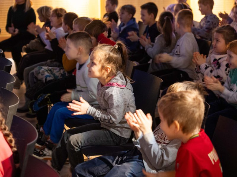 Dzieci na widowni klaszczą podczas występu; w centrum dziewczynka w szarej bluzie siedzi w rzędzie krzeseł