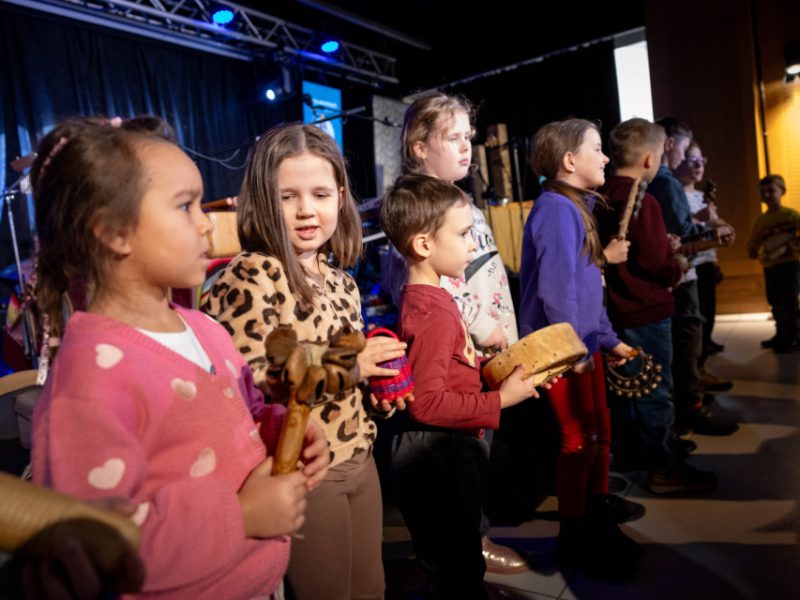 Rząd dzieci stoi na scenie z instrumentami w rękach (grzechotki, bębenek, tamburyn) podczas zajęć edukacyjnych